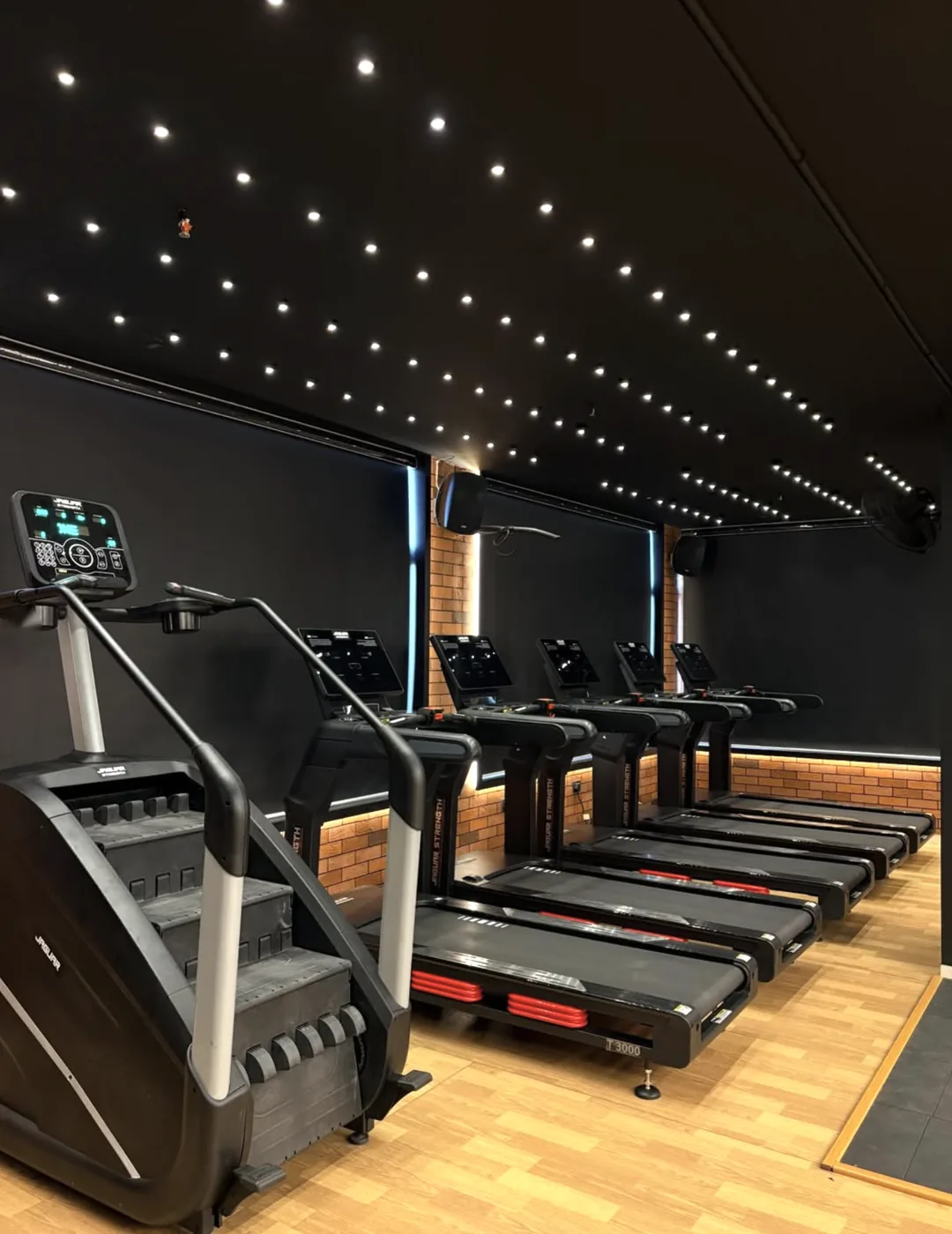 Row of treadmills with a stair climber in the foreground, starry ceiling lights and brick-effect wall